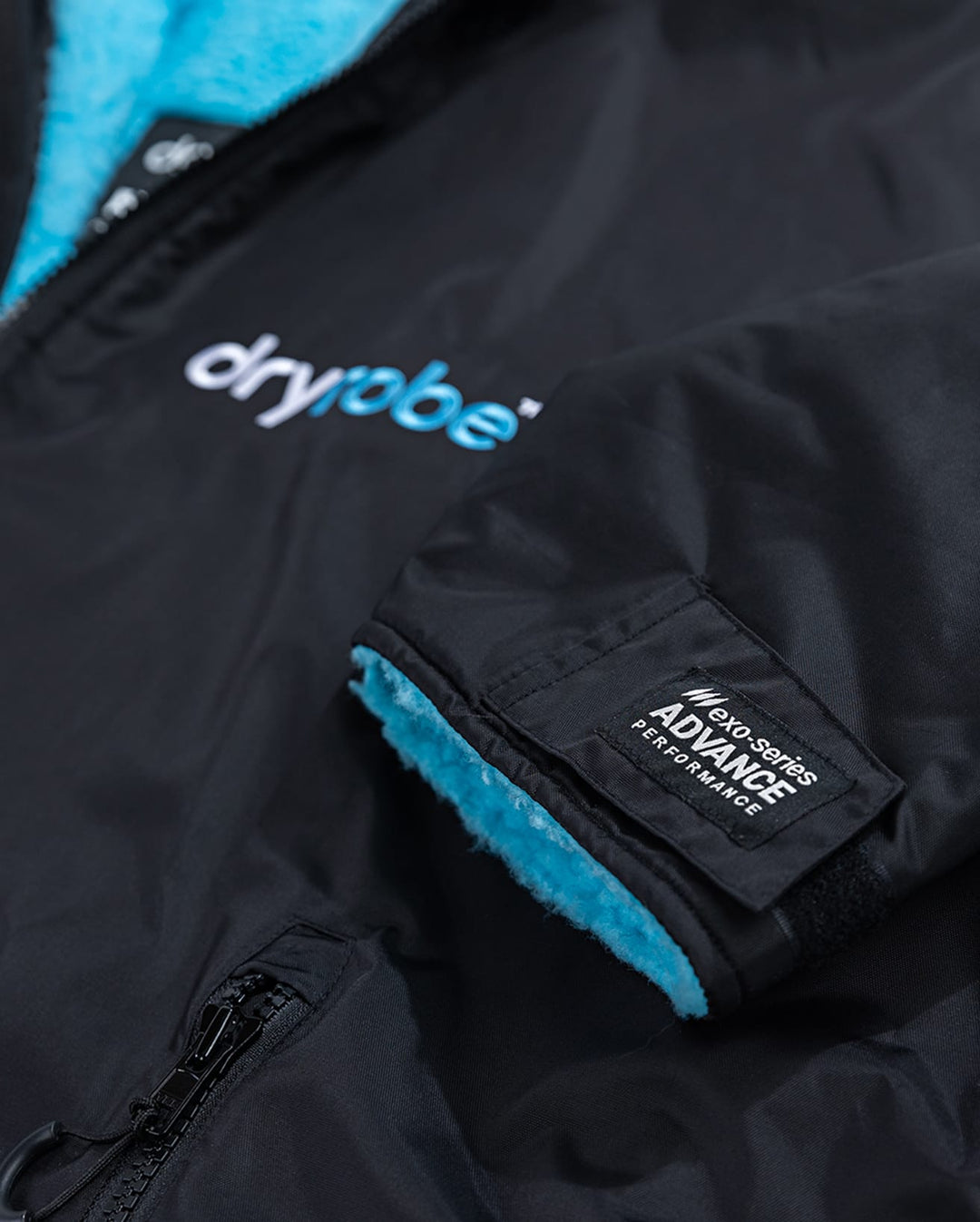 Nahaufnahme des Dryrobe Long Sleeve Schwarz Blau von Dryrobe, ein schwarzer Wickelponcho mit blauem Fleecefutter, mit dem Dryrobe-Logo auf der Brust, einem ADVANCE PERFORMANCE-Label auf dem Ärmel und einem teilweise sichtbaren Reißverschluss.