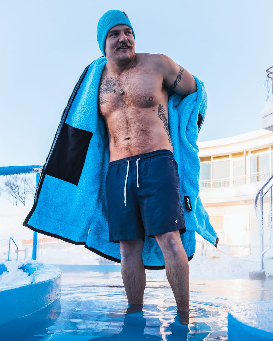 Ein Mann steht im Freien in einer verschneiten Gegend und trägt eine Badekappe, blaue Shorts und das Dryrobe Long Sleeve Schwarz Blau von Dryrobe. Im Hintergrund sind Gebäude und Geländer zu sehen, während er in einem Wasserbecken steht.