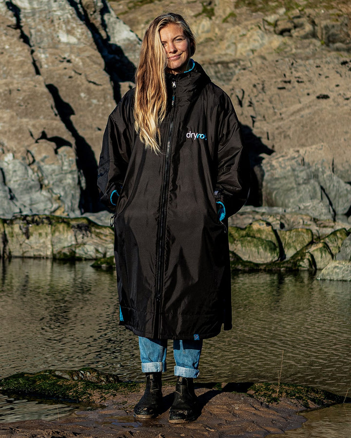 Eine Person steht im Freien an einer felsigen Küste und trägt den Dryrobe Long Sleeve Schwarz Blau von Dryrobe - einen wetterfester Umziehponcho - mit hochgekrempelten Jeans und schwarzen Stiefeln. Die Person hat lange Haare und lächelt leicht in die Kamera.
