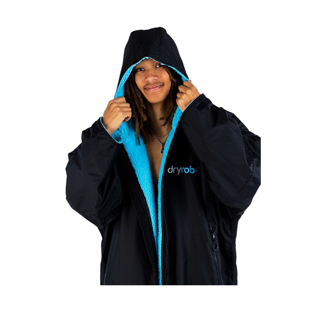 Eine Person, die ein Dryrobe Long Sleeve Schwarz Blau von Dryrobe trägt, hält den schwarz-blauen Mantel an der Brust offen und lächelt in die Kamera, wobei das "dryrobe"-Logo auf diesem wetterfesten Umziehponcho sichtbar ist.