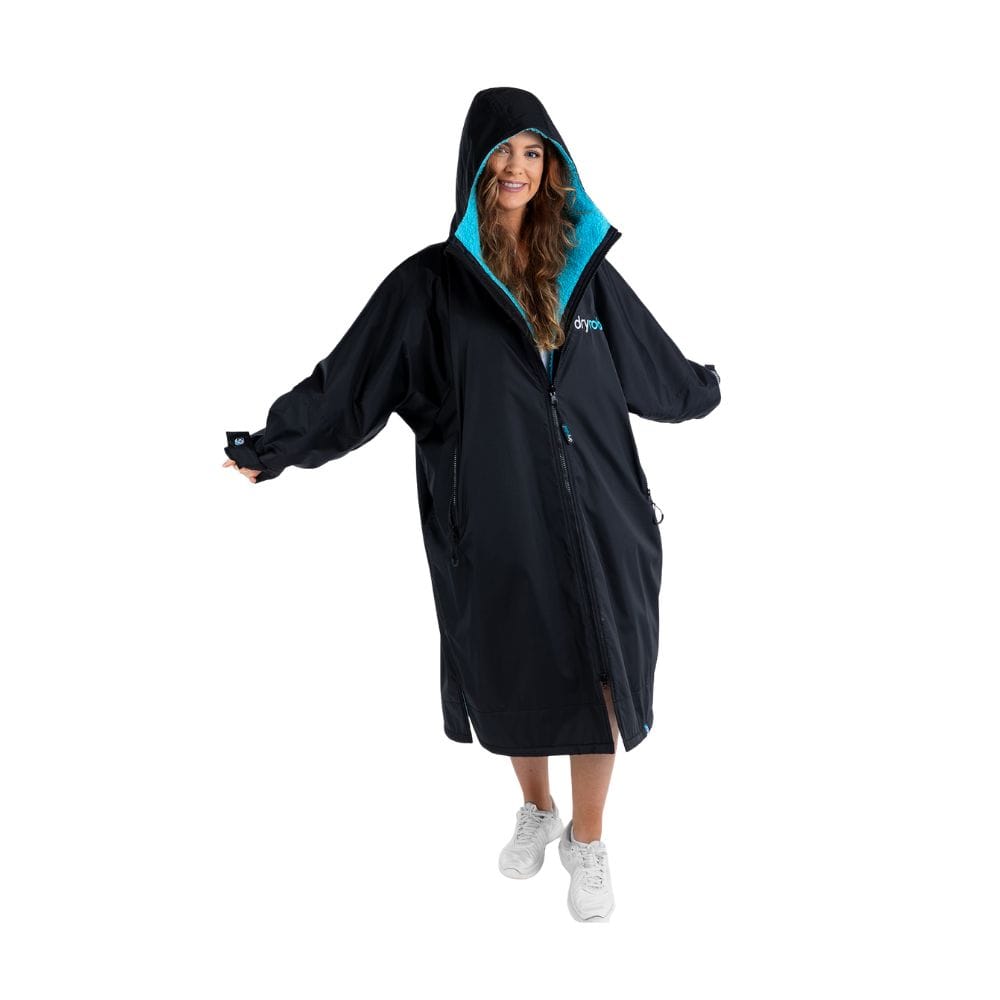 Eine Frau steht lächelnd mit ausgestreckten Armen da und trägt den Dryrobe Long Sleeve Schwarz Blau von Dryrobe - einen wetterfester Umziehponcho mit hellblauem Futter und weißen Turnschuhen - vor einem schlichten weißen Hintergrund.