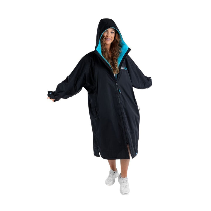 Eine Frau steht lächelnd mit ausgestreckten Armen da und trägt den Dryrobe Long Sleeve Schwarz Blau von Dryrobe - einen wetterfester Umziehponcho mit hellblauem Futter und weißen Turnschuhen - vor einem schlichten weißen Hintergrund.