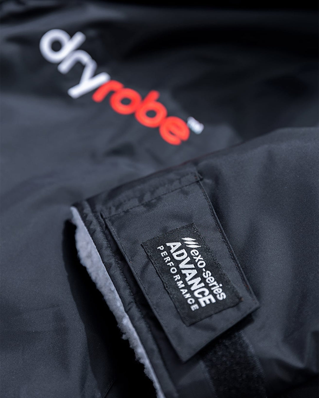 Eine Nahaufnahme der Dryrobe Long Sleeve Schwarz Grau Jacke mit dem weiß-roten Dryrobe-Logo und einem rechteckigen Etikett mit der Aufschrift eco series ADVANCE PERFORMANCE, das recyceltes Fleecefutter für mehr Nachhaltigkeit enthält.