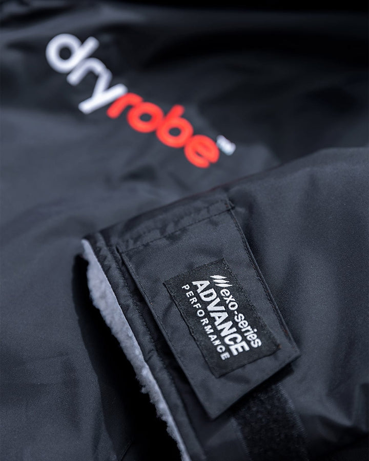 Eine Nahaufnahme der Dryrobe Long Sleeve Schwarz Grau Jacke mit dem weiß-roten Dryrobe-Logo und einem rechteckigen Etikett mit der Aufschrift eco series ADVANCE PERFORMANCE, das recyceltes Fleecefutter für mehr Nachhaltigkeit enthält.