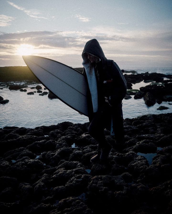 Eine Person, die das Dryrobe Long Sleeve Schwarz Grau von Dryrobe trägt, steht bei Sonnenuntergang mit einem weißen Surfbrett am felsigen Ufer. Der bewölkte Himmel und das Meer spiegeln die untergehende Sonne. Die Jacke verfügt über recyceltes Fleecefutter für Wärme.