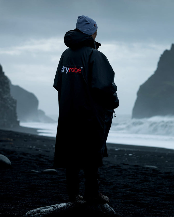 Eine Person, die ein Dryrobe Long Sleeve Schwarz Grau von Dryrobe trägt, steht an einem dunklen, felsigen Strand mit Blick auf das Meer, während die Wellen unter einem bewölkten Himmel gegen große Felsen schlagen.