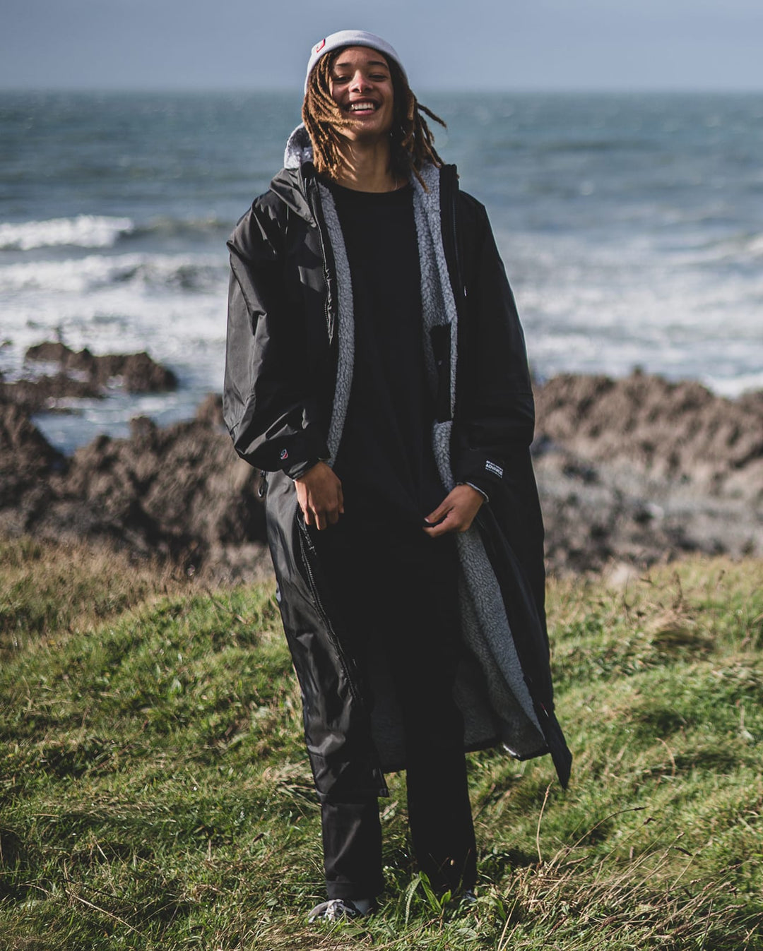 Eine Person, die ein Dryrobe Long Sleeve Schwarz Grau von Dryrobe trägt, steht auf einem grasbewachsenen Gelände in der Nähe einer felsigen Küste, während im Hintergrund unter einem wolkenverhangenen Himmel Meereswellen rauschen.
