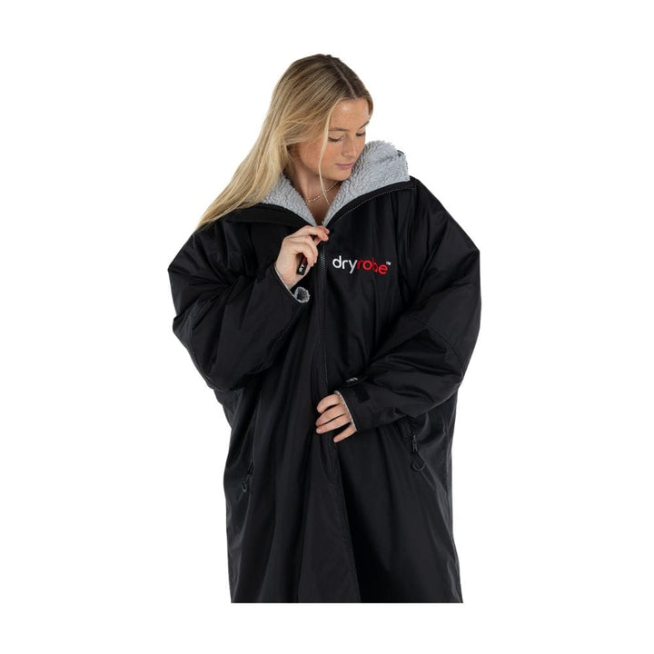 Eine Person mit langen blonden Haaren trägt ein teilweise ausgezogenes Dryrobe Long Sleeve Schwarz Grau von Dryrobe, mit recyceltes Fleecefutter, und schaut nach unten. Das Dryrobe-Logo ist auf der Vorderseite sichtbar.