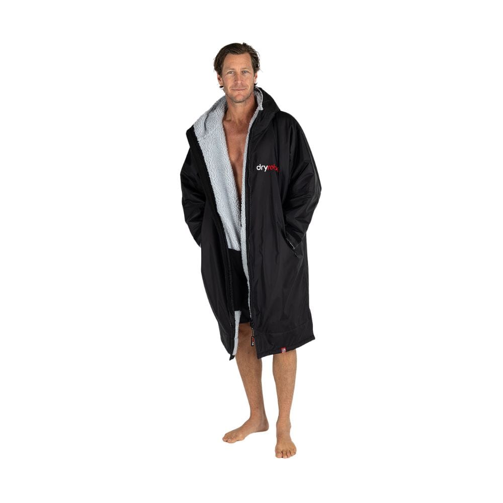 Ein Mann steht barfuß in einem Dryrobe Long Sleeve Schwarz Grau von Dryrobe, das geöffnet ist, um schwarze Shorts und einen Teil seiner nackten Brust zu zeigen. Er ist vor einem schlichten weißen Hintergrund zu sehen.