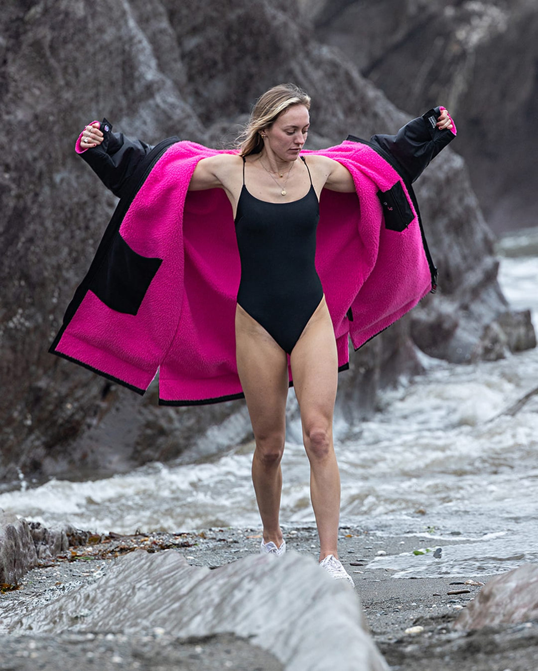 Eine Frau in einem schwarzen Badeanzug und weißen Schuhen zieht die Dryrobe Long Sleeve Schwarz Rosa von Dryrobe an, während sie auf Felsen in der Nähe von rauem, grauem Wasser mit felsigen Klippen hinter ihr steht.