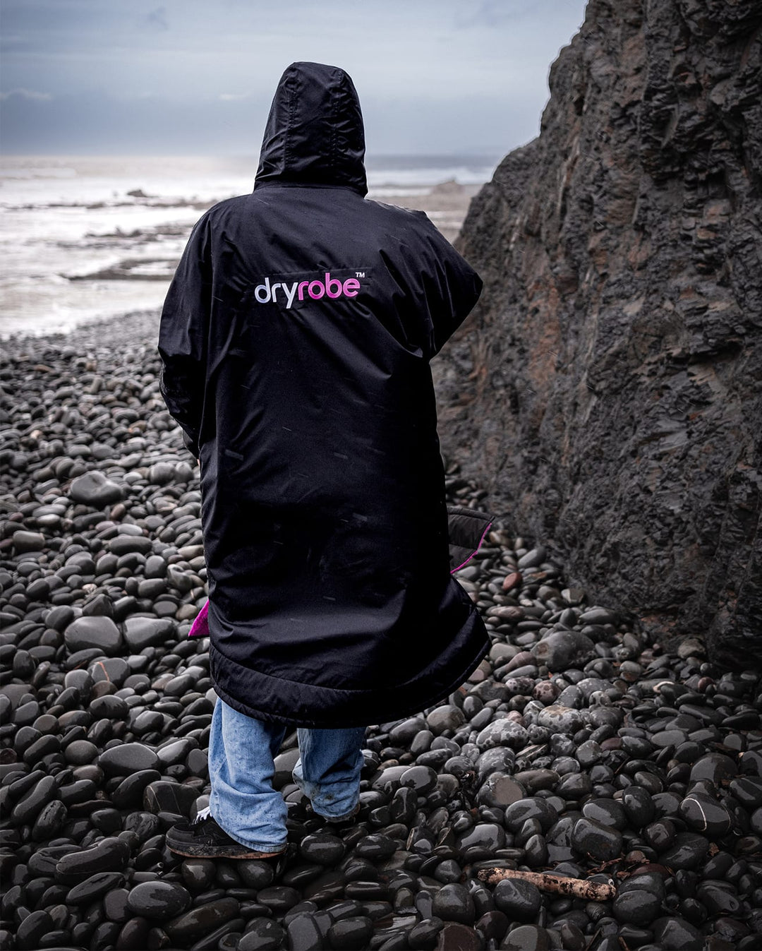 Eine Person, die ein Dryrobe Long Sleeve Schwarz Rosa von Dryrobe trägt, steht an einem felsigen Strand mit Blick auf den Ozean, mit großen Felsen und bewölktem Himmel im Hintergrund.