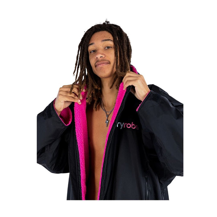 Eine Person mit langen Dreadlocks trägt das Dryrobe Long Sleeve Schwarz Rosa von Dryrobe, wobei sie das leuchtend rosafarbene Innenfutter zur Schau stellt und den Kragen wie eine mobile Umkleide vor einem weißen Hintergrund offen hält.