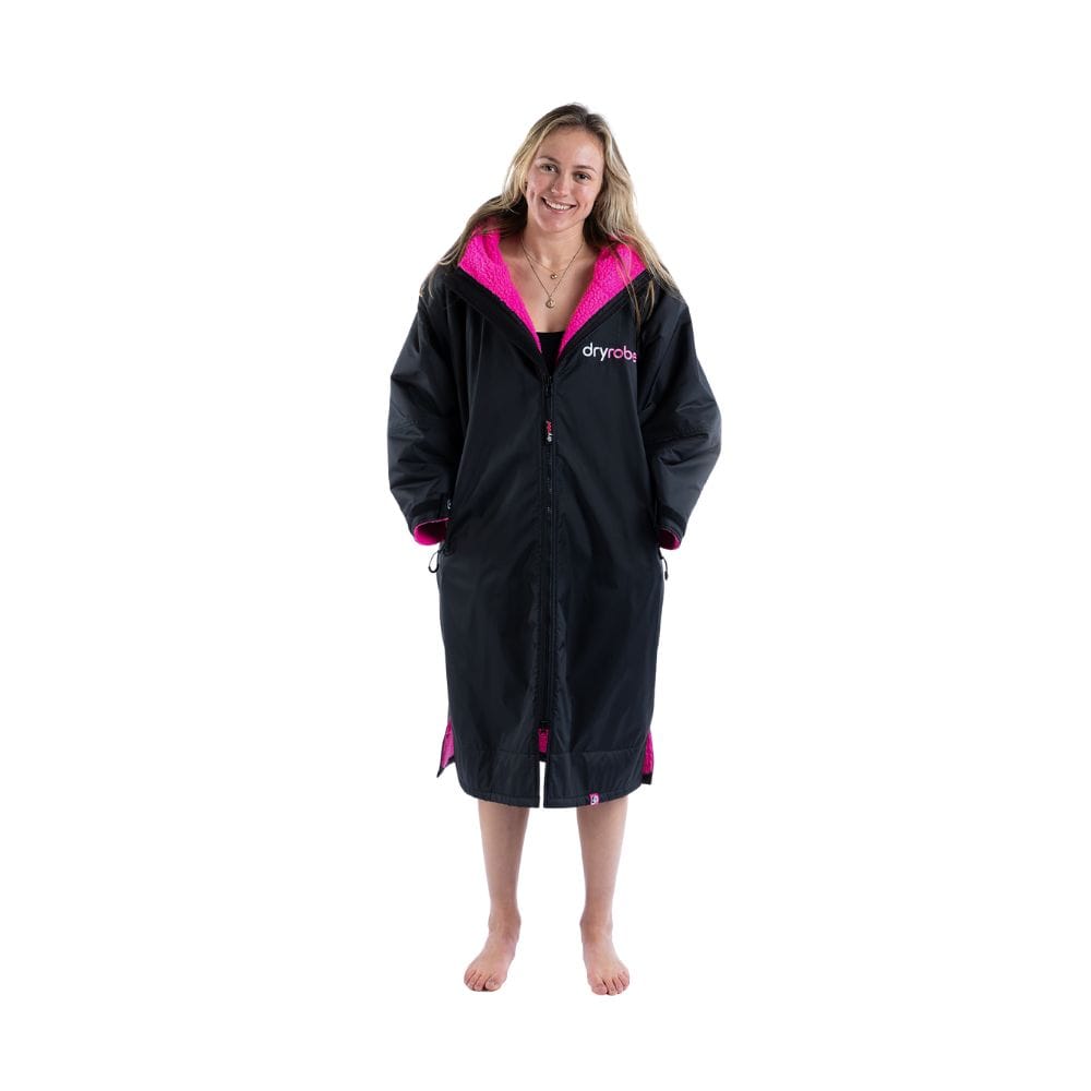 Eine Person steht barfuß, schaut nach vorne und lächelt. Sie trägt den Dryrobe Long Sleeve Schwarz Rosa von Dryrobe, einen schwarzen langen Mantel mit rosa Futter und Logo, der an einen wetterfesten Poncho vor einem schlichten weißen Hintergrund erinnert.