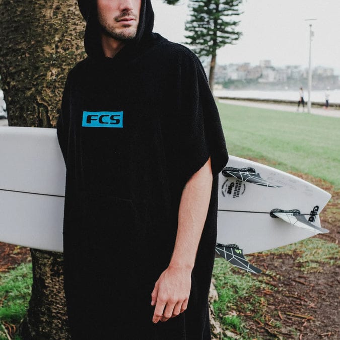 Eine Person, die den schwarzen FCS Surf Poncho (Handtuch-Frottee) 24er Edition von FCS trägt, steht draußen in der Nähe eines Baumes und hält ein weißes Surfbrett mit schwarzen Flossen. Im Hintergrund sind Gras, Bäume und entfernte Gebäude zu sehen.