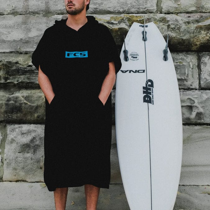 Eine Person, die den FCS Surf Poncho (Handtuch-Frottee) 24er Edition trägt, steht an einer Steinmauer, die Hände in den Taschen, neben einem weißen, aufrecht stehenden Surfbrett.