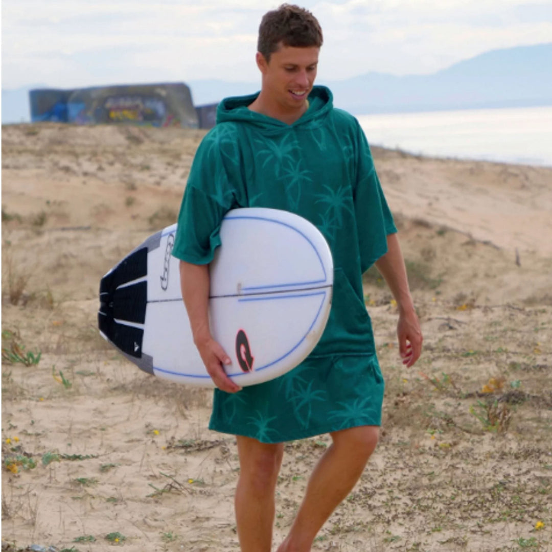 Ein Surfer trägt den MADNESS Change Robe Poncho Unisize Coconut von Madness mit Palmenmuster und grüner Farbe am Strand, hält ein weißes Surfbrett unter dem Arm. Im Hintergrund sind das Meer und ferne Hügel sichtbar.