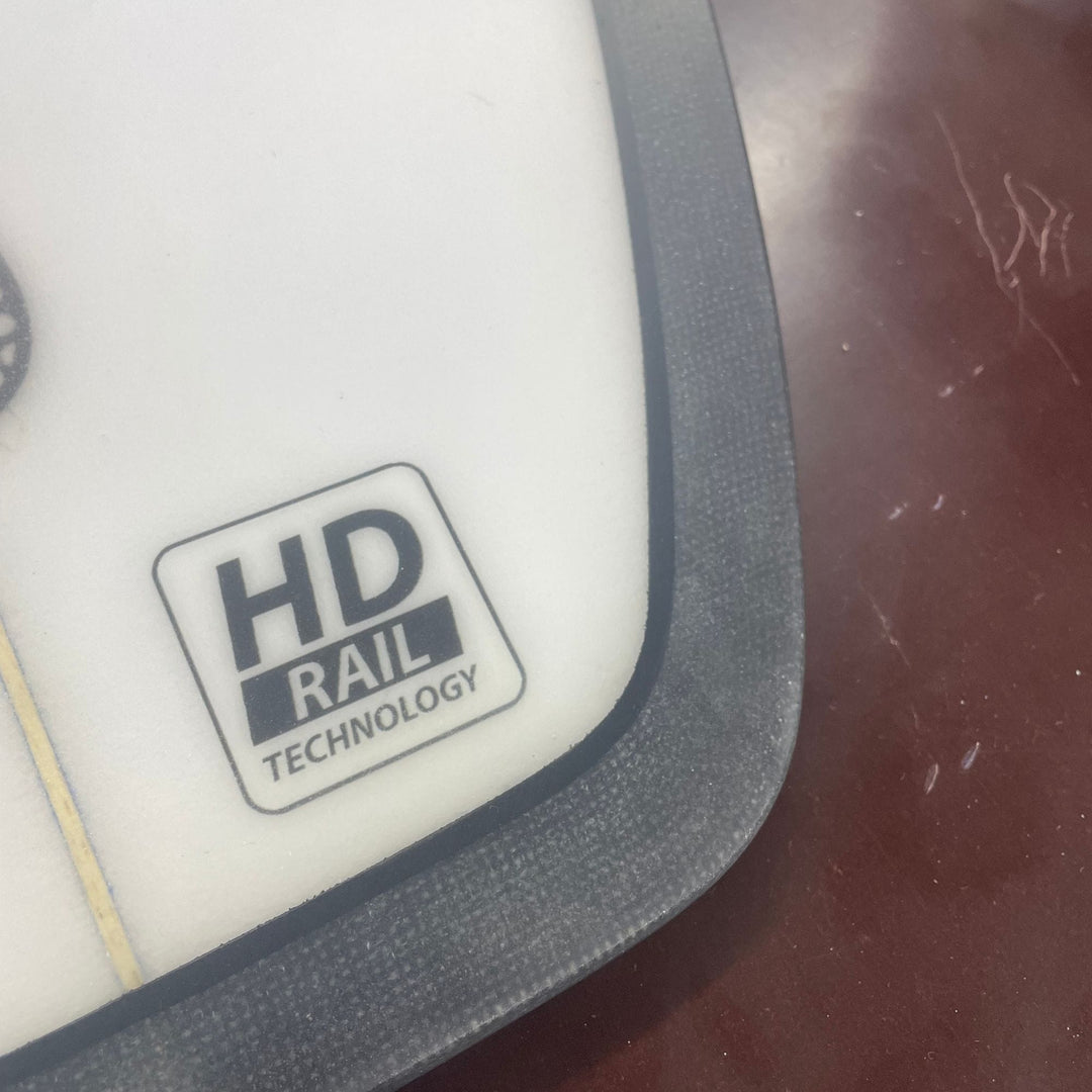 Die Nahaufnahme zeigt eine weiße Oberfläche mit schwarzem Rand auf dem Delight Alliance - Hoodoo Swallow riversurfboard, das mit einem Etikett versehen ist und sich vor einem glänzenden, dunkelrotbraunen Hintergrund mit Kratzern abhebt, die auf eine robuste Konstruktion hindeuten.