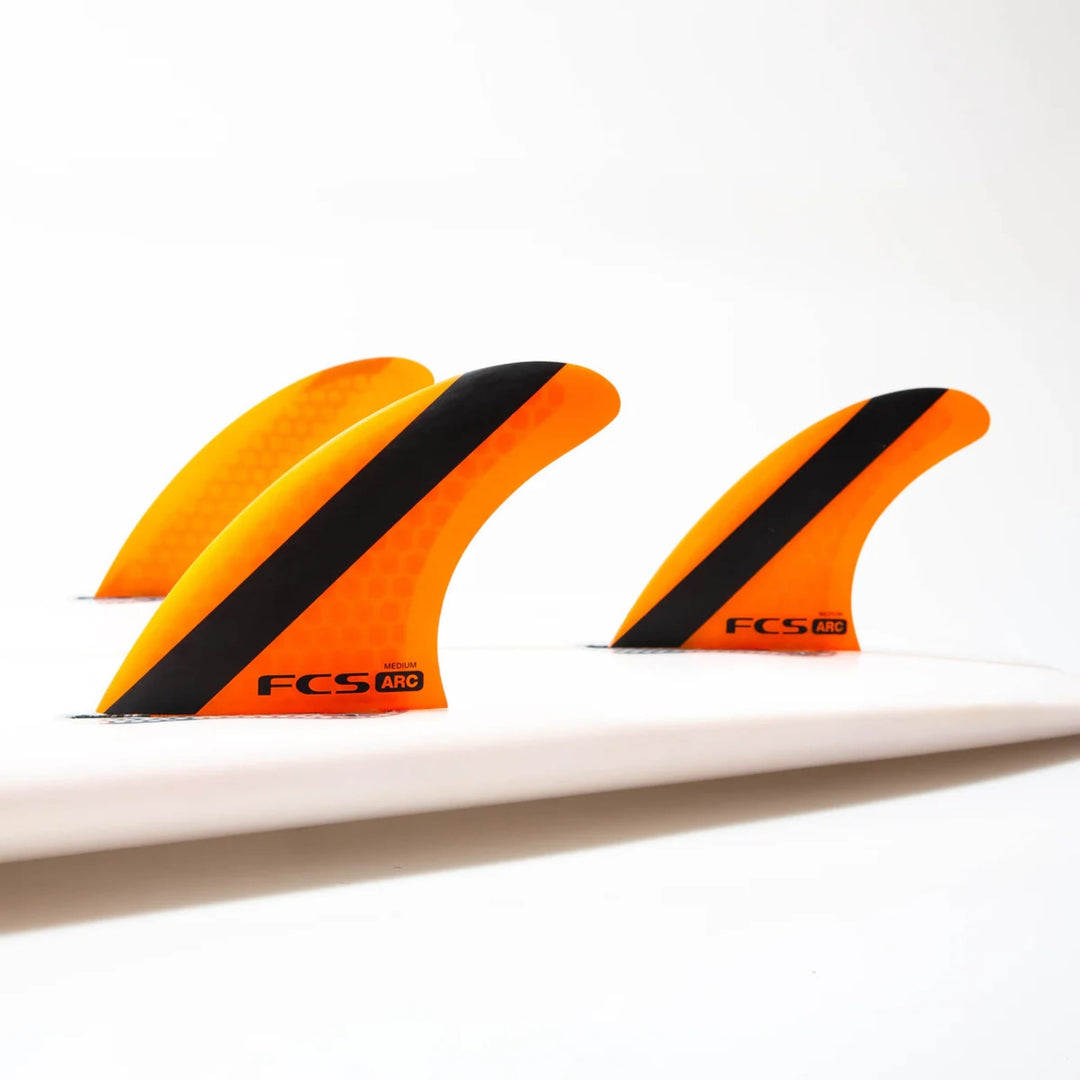 Drei leuchtend orangefarbene FCS 1 ARC Tri Fin Set Surfboard-Flossen mit schwarzen Streifen sind auf einem weißen Brett befestigt, das auf einem einfarbigen Hintergrund abgebildet ist. Hergestellt von FCS, verwenden diese Flossen Performance Core Technology für erweiterte Surfleistung.