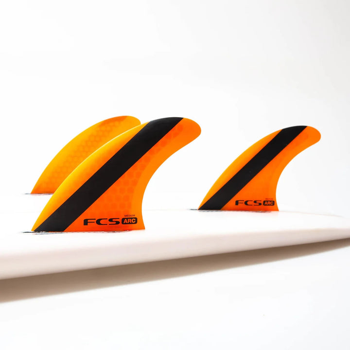 Drei leuchtend orangefarbene FCS 1 ARC Tri Fin Set Surfboard-Flossen mit schwarzen Streifen sind auf einem weißen Brett befestigt, das auf einem einfarbigen Hintergrund abgebildet ist. Hergestellt von FCS, verwenden diese Flossen Performance Core Technology für erweiterte Surfleistung.