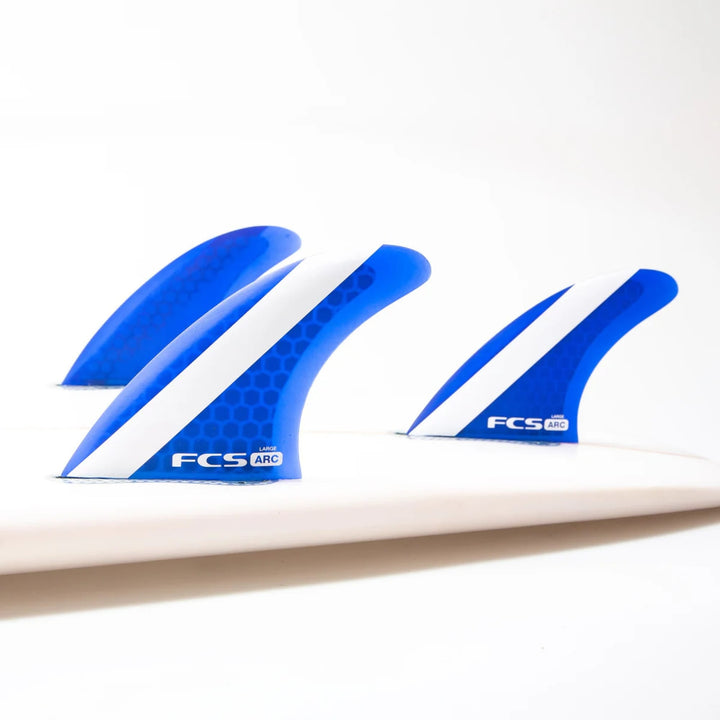 Nahaufnahme von drei blauen und weißen FCS 1 ARC Tri Fin Set Surfboard-Flossen von FCS mit Performance Core Technology, die auf einem weißen Surfbrett vor einem schlichten weißen Hintergrund angebracht sind.