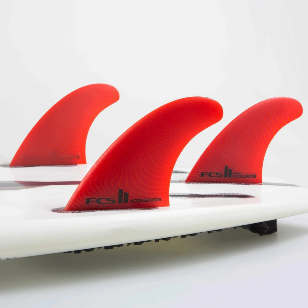 Eine Nahaufnahme von drei FCS 2 Accelerator Neo Glass Red Tri Fins von FCS, beschriftet mit "FCS II Accelerator", montiert auf einem weißen Surfbrett für Performance Shortboards, vor einem schlichten weißen Hintergrund.