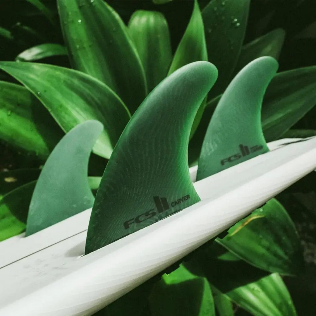 Nahaufnahme von drei grünen FCS 2 Carver Neo Glass Sage Tri Fins von FCS, die auf einem weißen Surfbrett vor üppigen grünen Blättern befestigt sind - für nachhaltige Leistung bei jeder Fahrt.