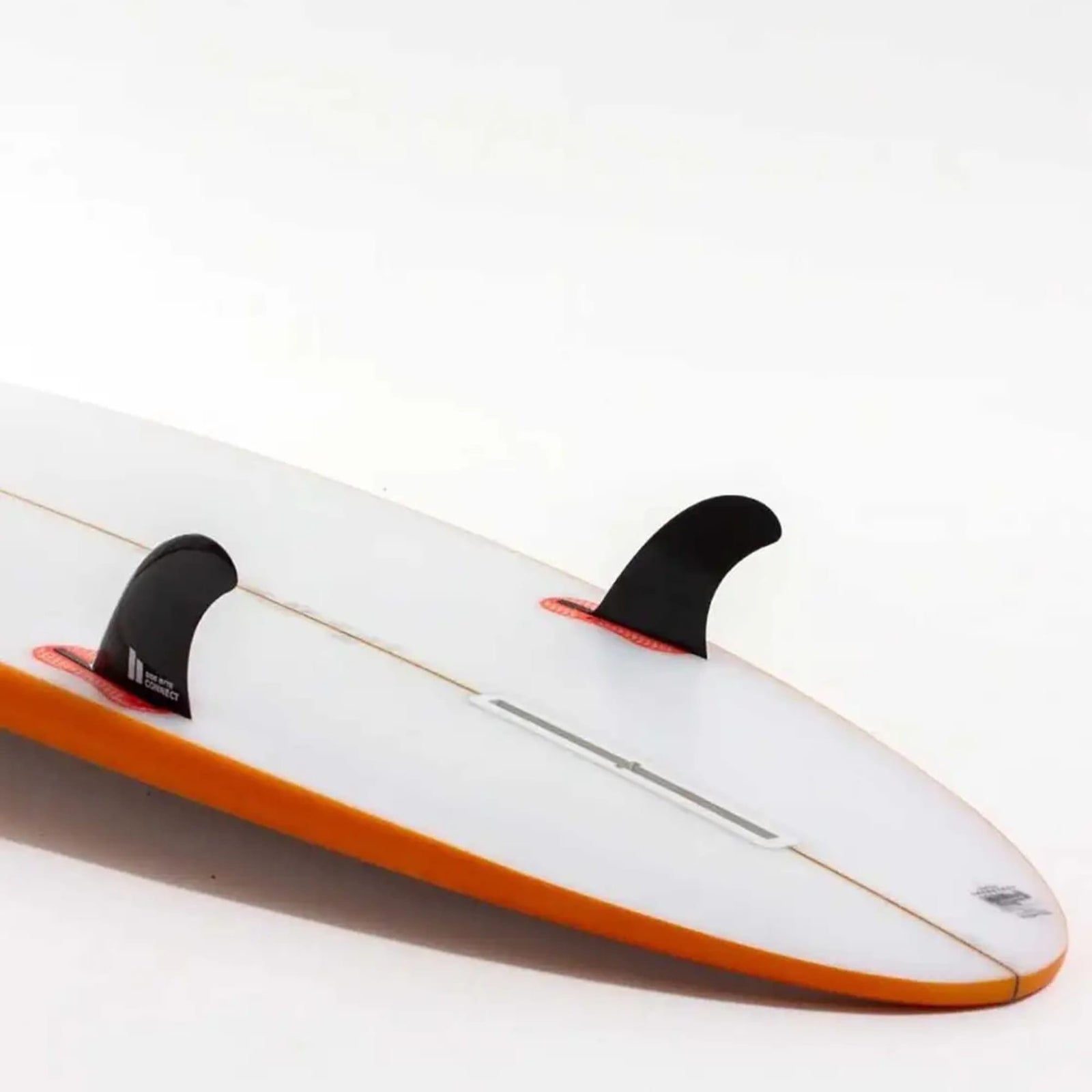 Eine Nahaufnahme eines weißen Surfbretts mit orangefarbenem Rand zeigt zwei FCS 2 Connect Black Small Quad Rear Side Byte Fins von FCS und eine leere Finnenbox, die ein mögliches Quad-Setup vor einem schlichten weißen Hintergrund hervorhebt.