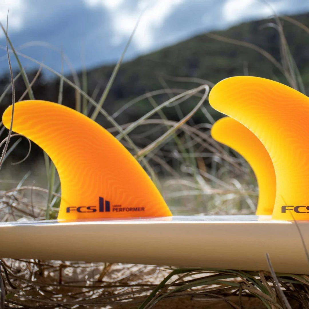 Eine Nahaufnahme von drei FCS 2 Performer Neo Glass Mango Tri Fins von FCS, die an einem weißen Surfbrett befestigt sind, mit hohem Gras und einer verschwommenen Landschaft im Hintergrund.