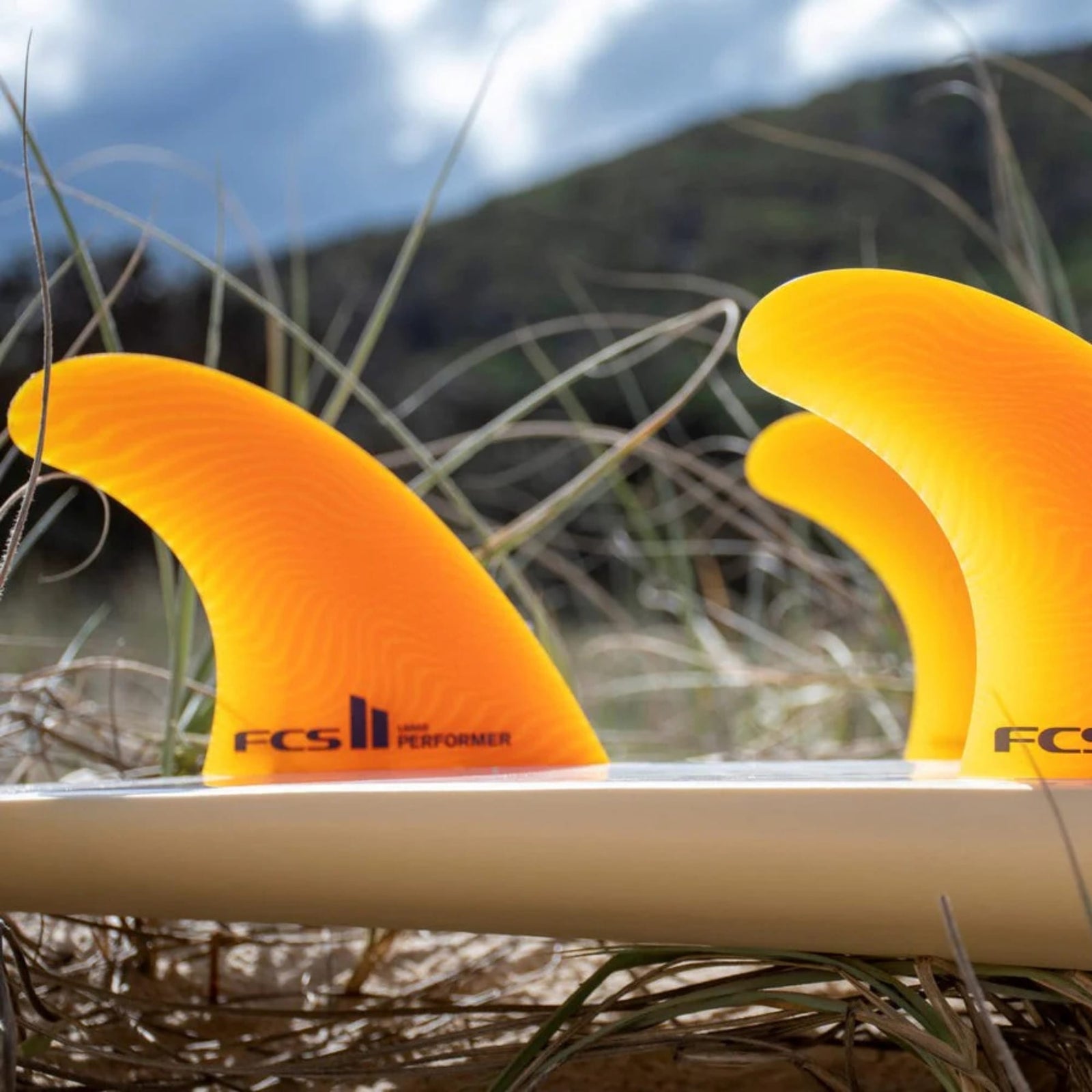 Eine Nahaufnahme von drei FCS 2 Performer Neo Glass Mango Tri Fins von FCS, die an einem weißen Surfbrett befestigt sind, mit hohem Gras und einer verschwommenen Landschaft im Hintergrund.
