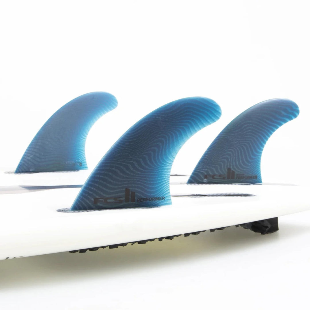 Drei FCS 2 Performer Neo Glass Pacific Tri Fins von FCS, in blau mit Wellenmuster, sind in einer Reihe auf einem weißen Surfbrett befestigt, von der Seite gesehen vor einem weißen Hintergrund.