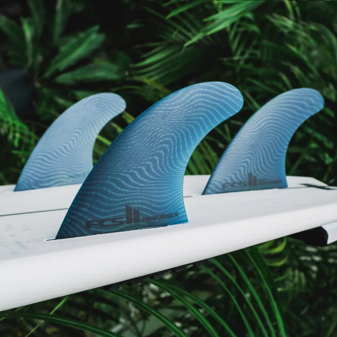 Nahaufnahme von drei blauen FCS 2 Performer Neo Glass Pacific Tri Fins von FCS auf einem weißen Surfbrett, die ein subtiles Wellenmuster und den Text "FCS II Performer" zeigen, mit verschwommenen grünen tropischen Blättern im Hintergrund.