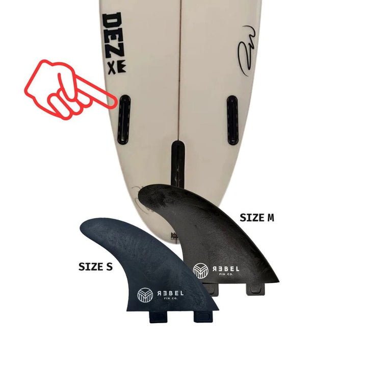 Ein Surfbrett mit zwei schwarzen Finnenkästen steht aufrecht. Davor befinden sich zwei abnehmbare Rebel Fin - einzelne Finne (Carbon) von Rebel Fin, beschriftet mit Größe S und Größe M. Ein rotes Handsymbol zeigt auf einen der Finnenkästen des Surfboards.