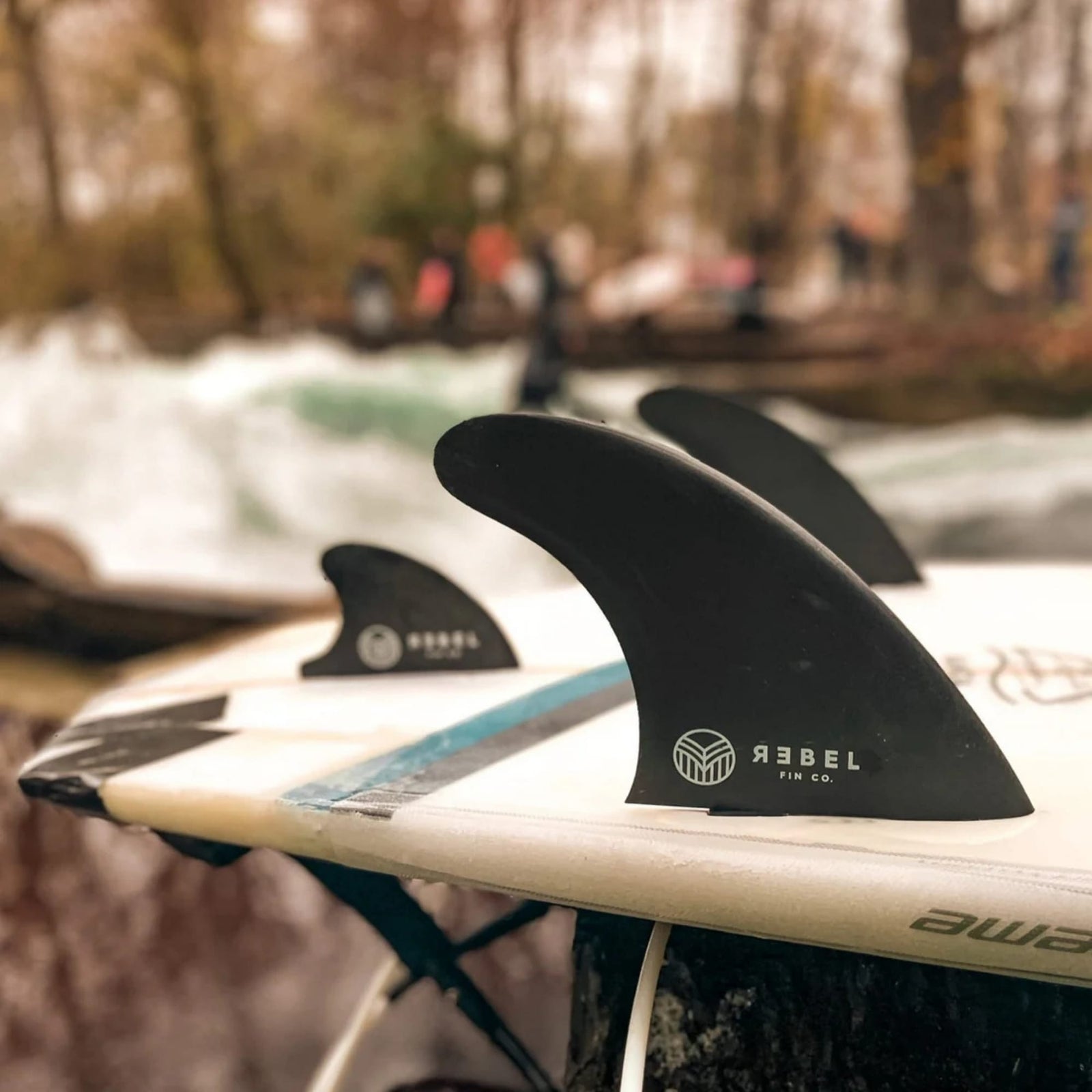 Nahaufnahme der Rebel Fin - Twin Set Carbon schwarz von Rebel Fin, montiert auf einem weißen Surfbrett im Freien in der Nähe eines Flusses, mit unscharfen Bäumen und Menschen im Hintergrund.