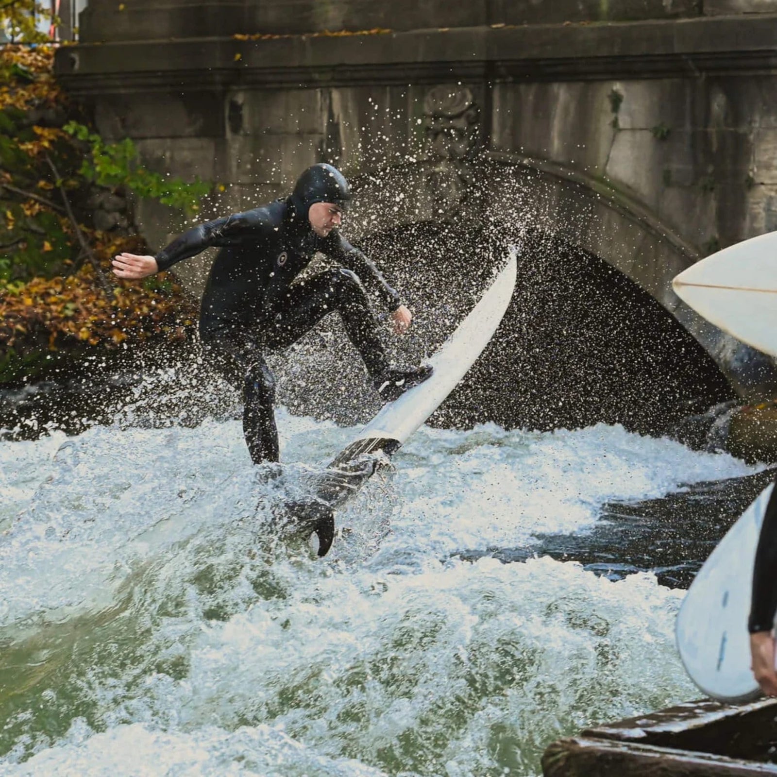 Ein Surfer im Neoprenanzug reitet auf einer stehenden Welle und balanciert auf einem Brett, während das Wasser spritzt. In der Nähe steht jemand mit der Rebel Fin - Rapid Fin Set FCS I - Carbon von Rebel Fin. Eine gewölbte Steinbrücke und Herbstbäume erscheinen im Hintergrund.