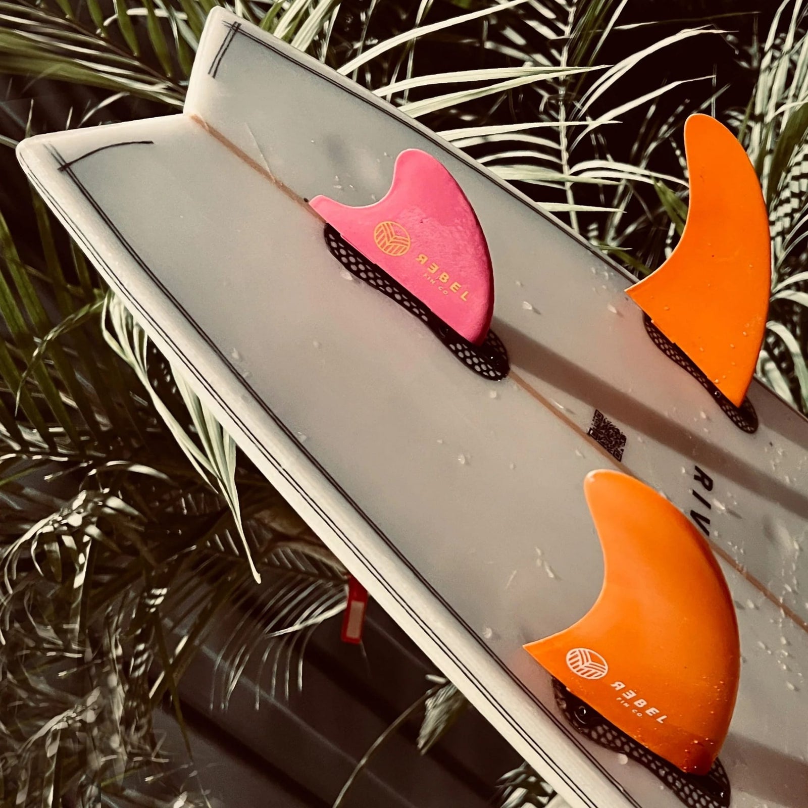 Nahaufnahme eines weißen Surfbretts mit dem Rebel Fin - Rapid Fin Set FCS I - Flex orange von Rebel Fin-zwei große orangefarbene Flexfinnen und eine kleine pinke Finne-stehen aufrecht vor grünen Palmenblättern und Holzplanken, ideal für stehende Wellen.