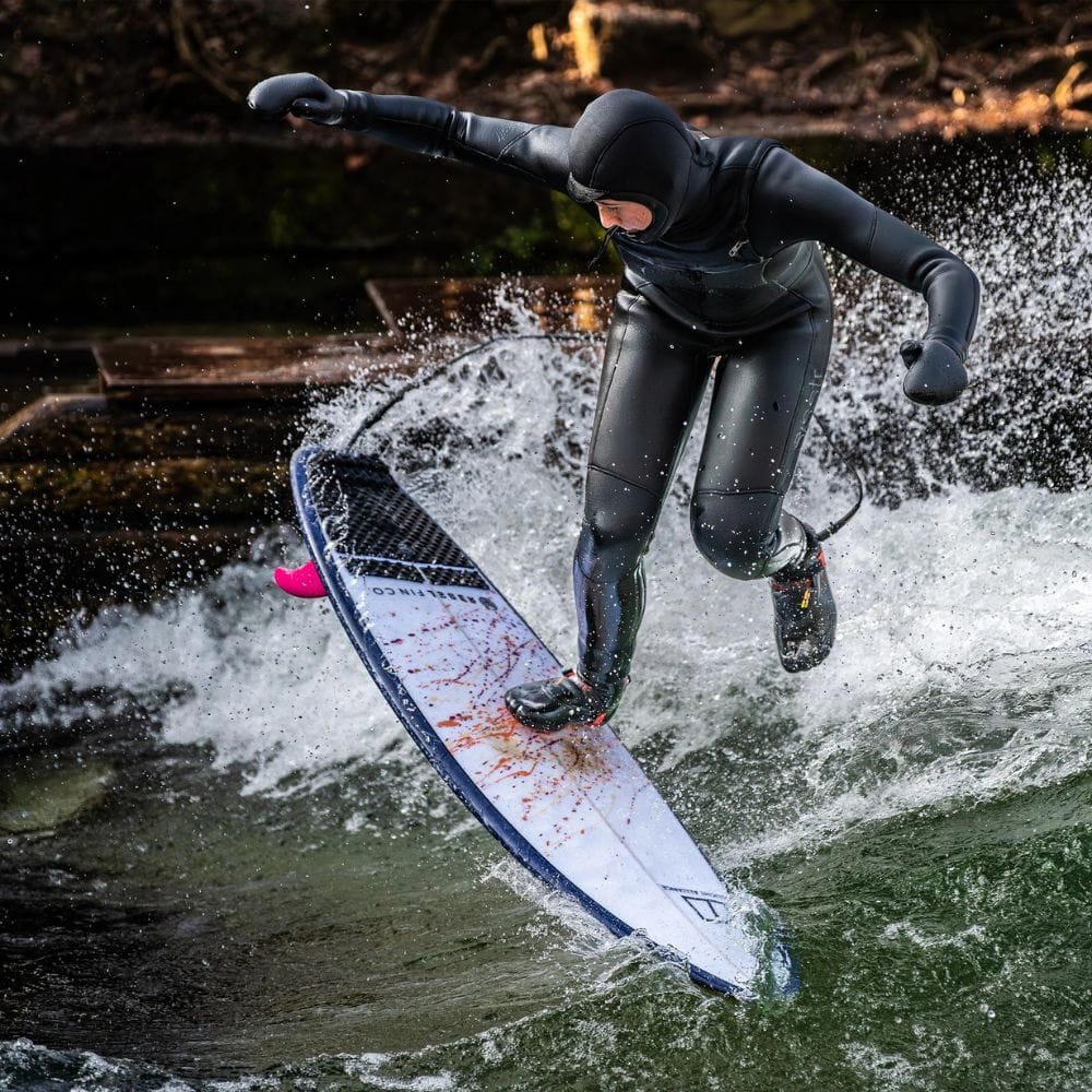 Bekleidet mit einem schwarzen Neoprenanzug und einer Kapuze balanciert ein Surfer auf einem Brett, das mit der Rebel Fin - Rapid Fin Set FCS I - Glasfaser pink von Rebel Fin ausgestattet ist, und gleitet mit erhobenem Arm durch die Wellen, während das Wasser um ihn herumspritzt.