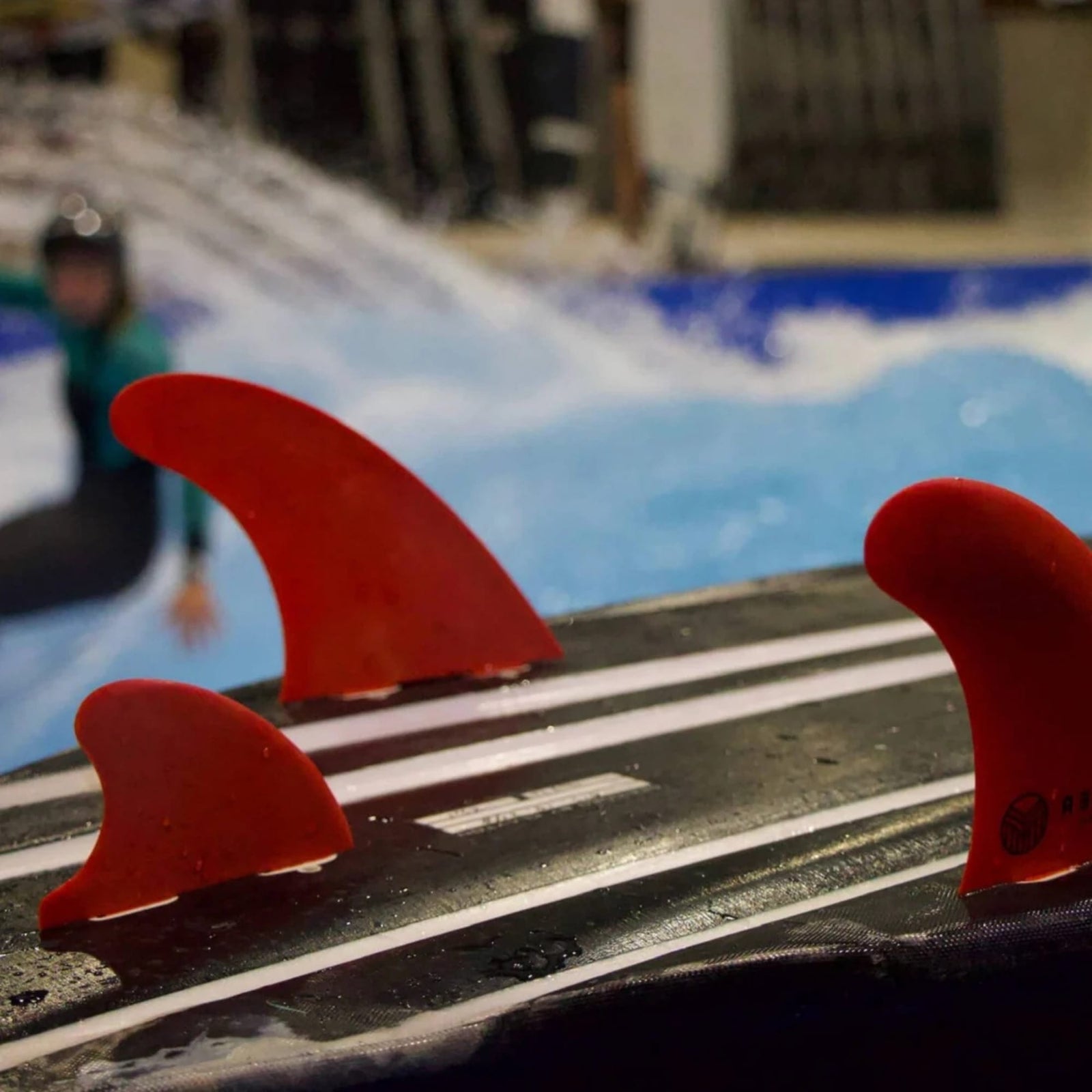 Nahaufnahme von drei Rebel X Driftwood - Fin Rapid Fin FCS I von Rebel Fin, hergestellt aus recycelter Glasfaser, im Fokus. Im unscharfen Hintergrund reitet ein behelmter Surfer auf einer Indoor-Welle, dahinter plätschert blaues Wasser.