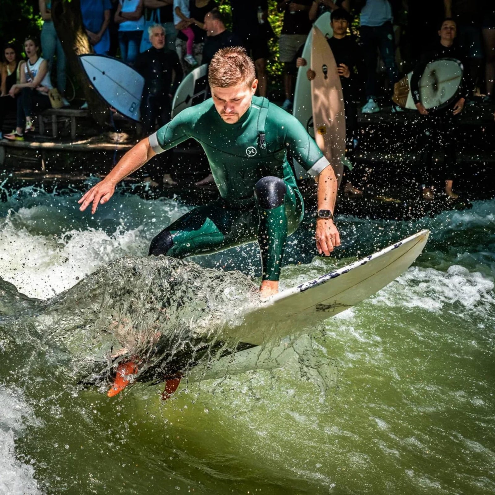 Ein Surfer in einem grünen Neoprenanzug reitet auf einer Welle und lehnt sich auf seinem mit dem Rapid Fin Set FCS I - Glasfaser rot von Rebel Fin ausgestatteten Brett nach vorne, während das Wasser spritzt und mehrere Personen mit Surfbrettern vom Ufer aus zusehen.