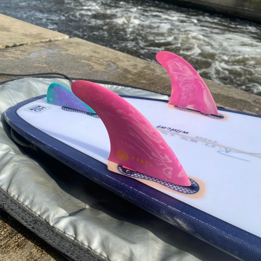 Eine Nahaufnahme eines Surfbretts mit dem Rebel Fin - Rapid Fin Set FCS I - Marble (zwei rosa Flossen, eine blaugrüne) von Rebel Fin, das mit der Flosse nach oben auf einer grauen Unterlage an einem Fluss liegt, bereit zum Fluss-Surfen.