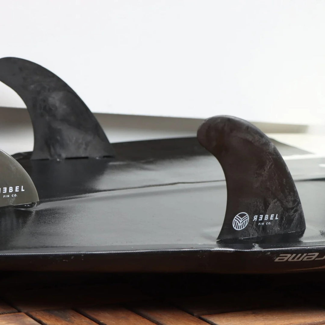 Nahaufnahme der Rebel Fin - Twin Set Carbon schwarz von Rebel Fin, die an einem Surfbrett befestigt ist, mit den Markenlogos. Das Surfbrett steht auf einer Holzfläche mit einer weißen Wand im Hintergrund.