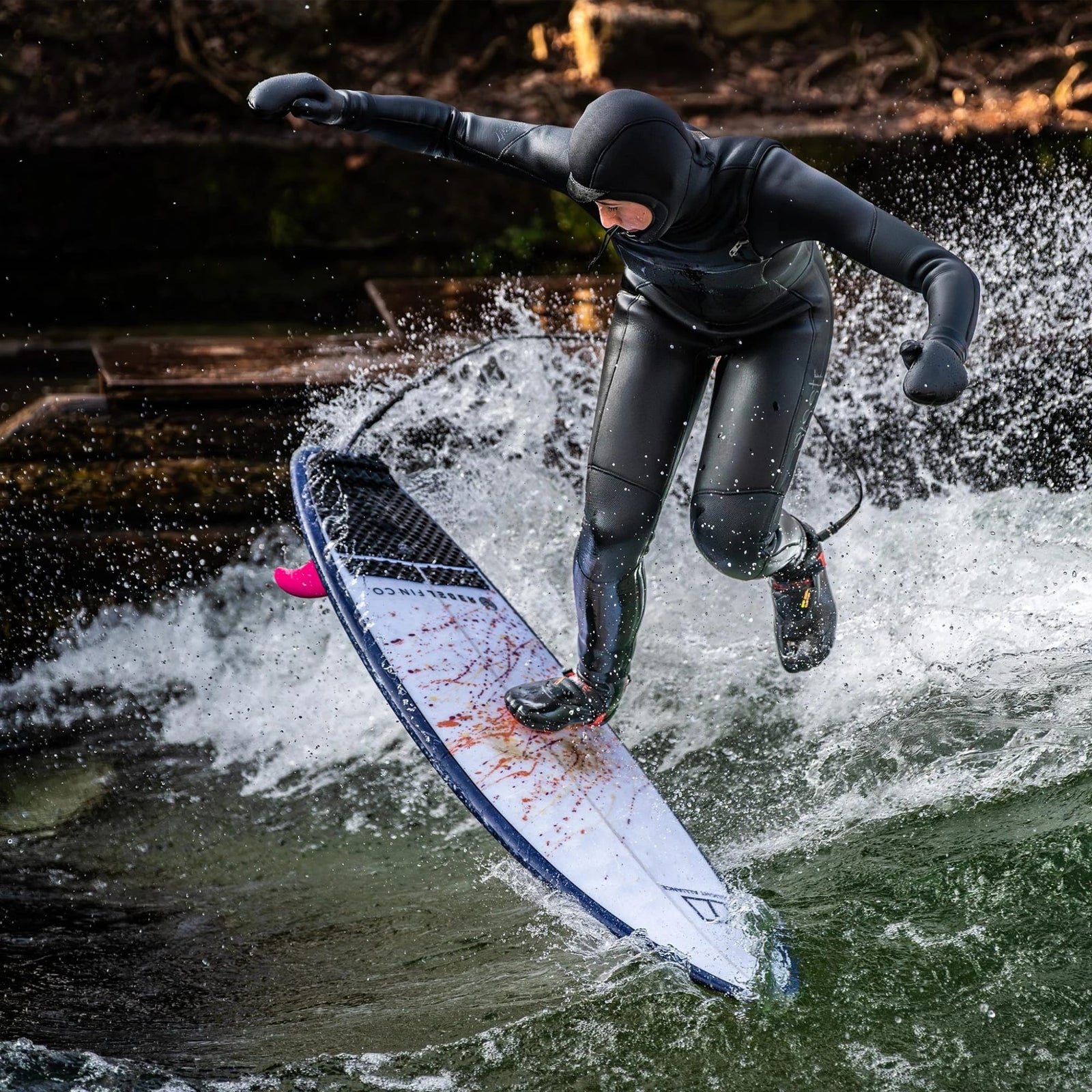 Ein Rapid Surfer in schwarzem Neoprenanzug und Kapuze verliert auf einer Welle das Gleichgewicht, das Wasser spritzt, während sein Surfbrett mit der Rebel Fin - Twin Set Glasfaser pink von Rebel Fin nach oben kippt.