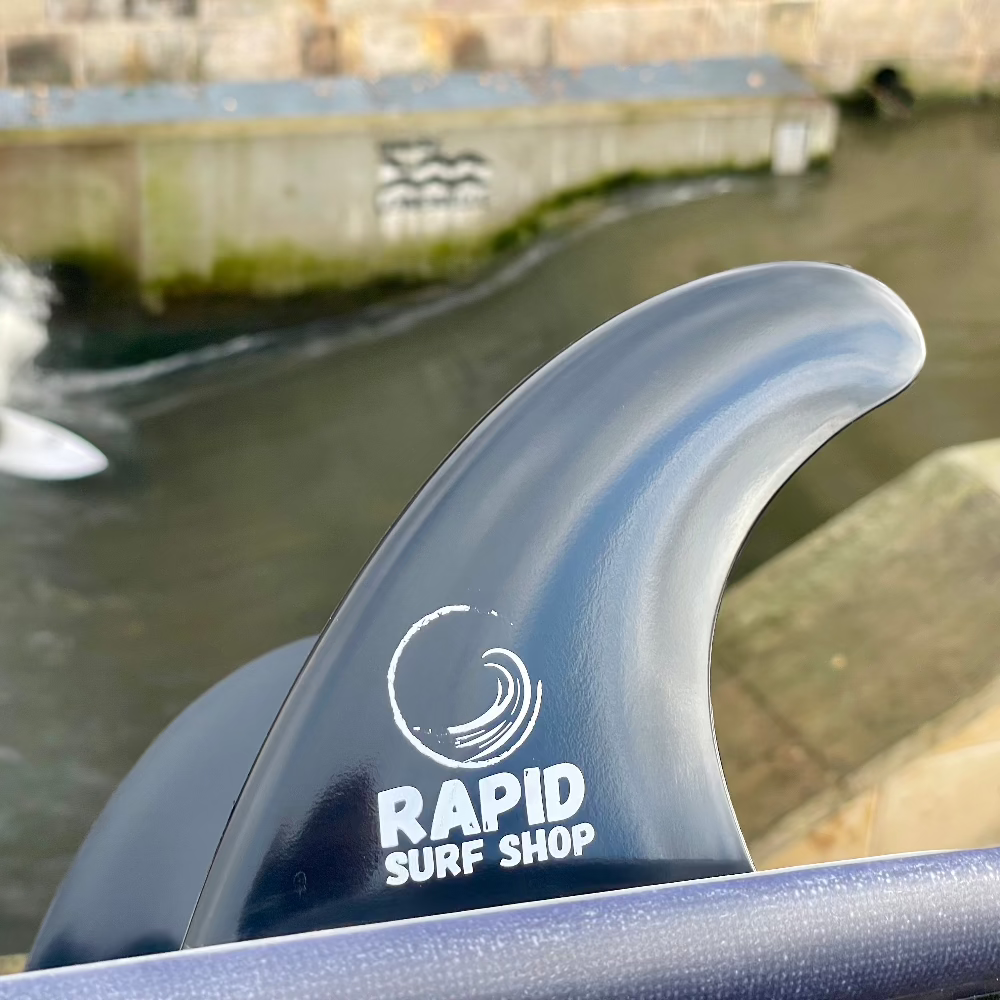 Nahaufnahme des Surfboards Finnen - Twin Set M FCSI von Rapid Surf Shop, mit dem weißen Logo des Shops auf einer schwarzen Finne. Im Hintergrund Wasser und eine mit Graffiti beschmierte Wand - ideal für Rapid Surfen Fans, die hochwertige Ersatzfinnen suchen.