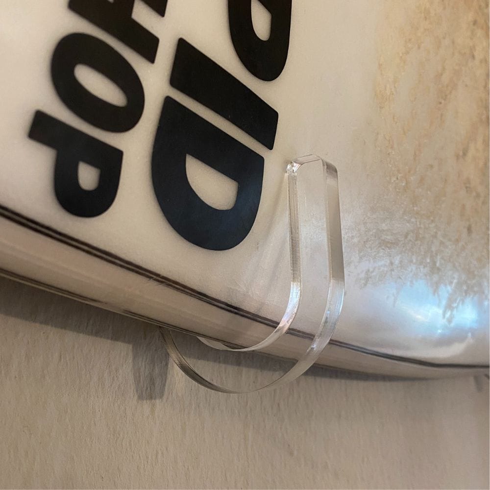 Eine Nahaufnahme der Rapid Surf Shop Surfboard Wandhalterung durchsichtig | B-Ware, die ein Surfbrett mit schwarzem Schriftzug auf weißem Hintergrund sicher hält.