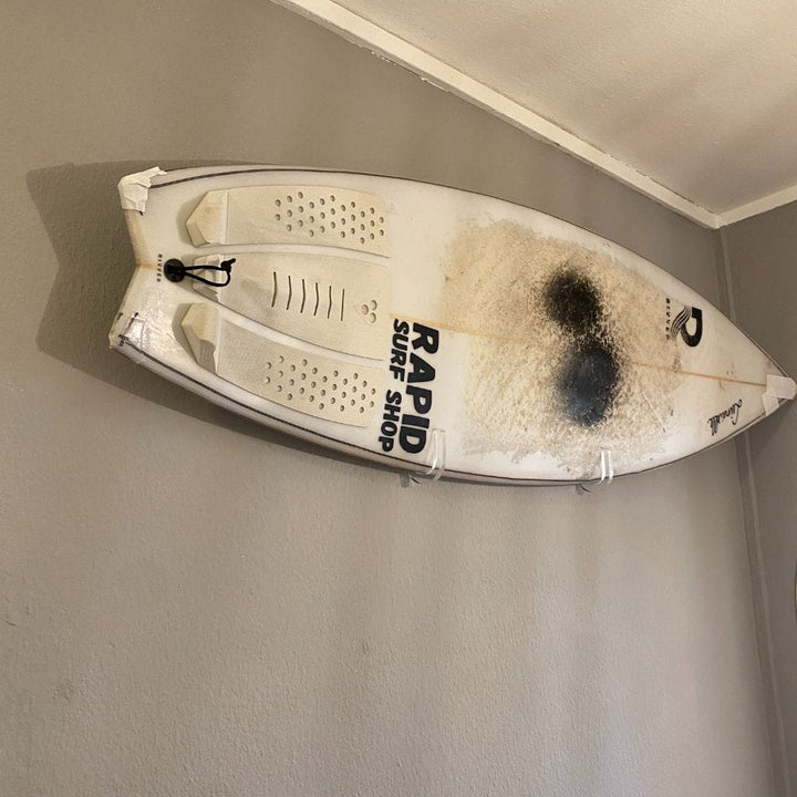 Ein weißes Surfbrett mit schwarzen Brandspuren und dem Schriftzug "Rapid Surf Shop" ist mit der transparenten Surfboard Wandhalterung durchsichtig | B-Ware von Rapid Surf Shop horizontal in Deckennähe an einer grauen Wand angebracht. Das Brett weist Gebrauchsspuren und Beschädigungen auf.