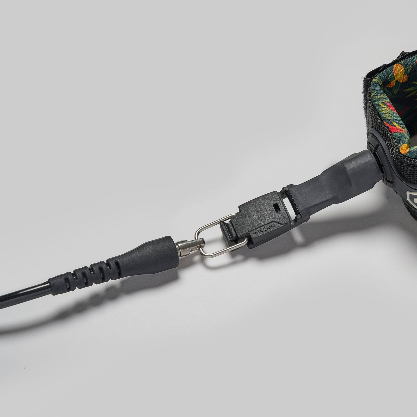 Eine Nahaufnahme der Edgeboards Surfboard Leash mit Quickrelease (Releash) zeigt das floral gemusterte Band, den Metallclip und die schwarze Kunststoffschnalle mit Schnellverschluss, die an die Leash-Systeme von Rapid Surfing erinnern, auf einem schlichten grauen Hintergrund.