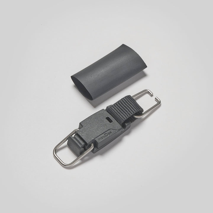 Eine schwarze Kunststoffschnalle mit Metallschlaufen und ein schwarzer Schrumpfschlauch - Hauptbestandteile der Magnetleash mit ReLeash Verschluss von Rapid Surf Shop - sind auf einem schlichten hellgrauen Hintergrund zu sehen.