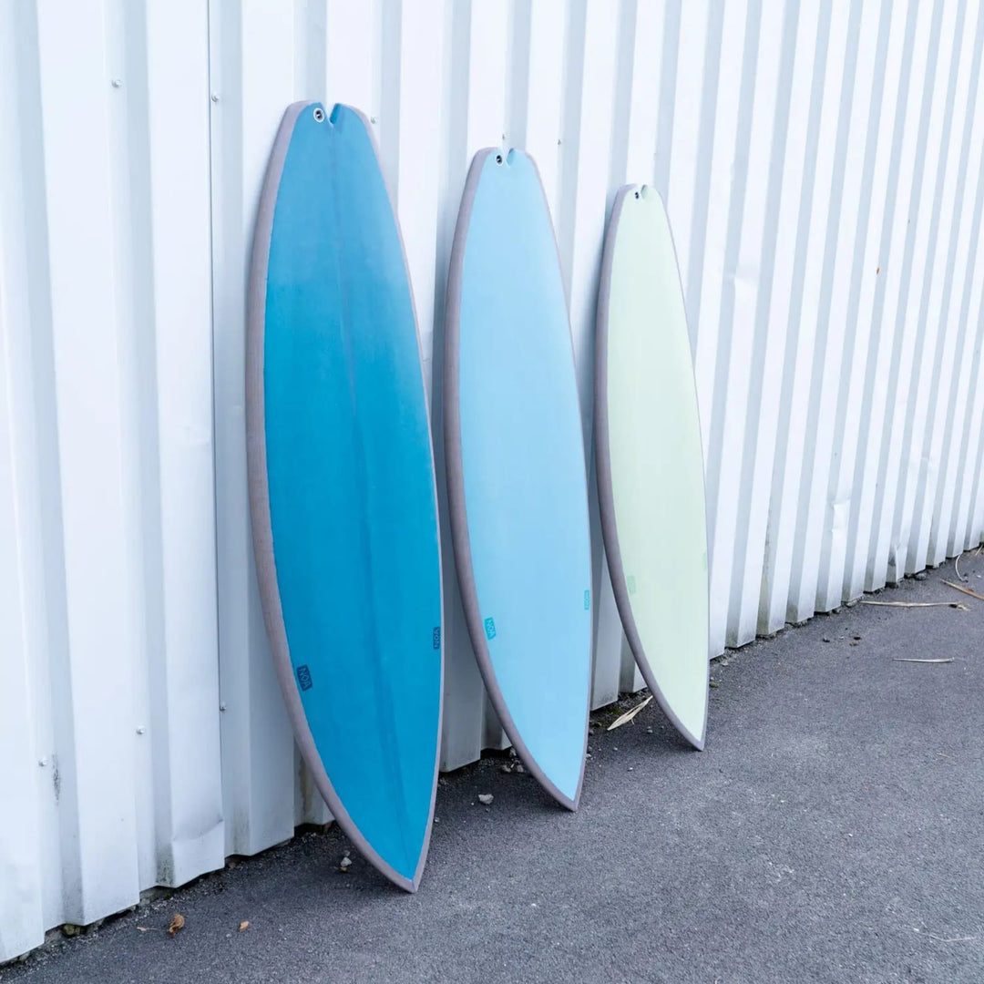 Drei NOA Surfboards - Bachforelle von NOA, in blauen und hellgrünen Farbtönen, stehen aufrecht vor einer weißen Wellblechwand auf einer grauen Betonfläche.