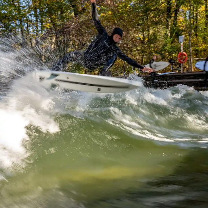 Eine Person in schwarzem Neoprenanzug und Kapuze reitet auf einer Flusswelle auf einem NOA Surfboards - Bachforelle von NOA, Wasser plätschert herum. Bäume mit grünen und gelben Blättern stehen im Hintergrund, während zwei Personen vom Flussufer aus zusehen.