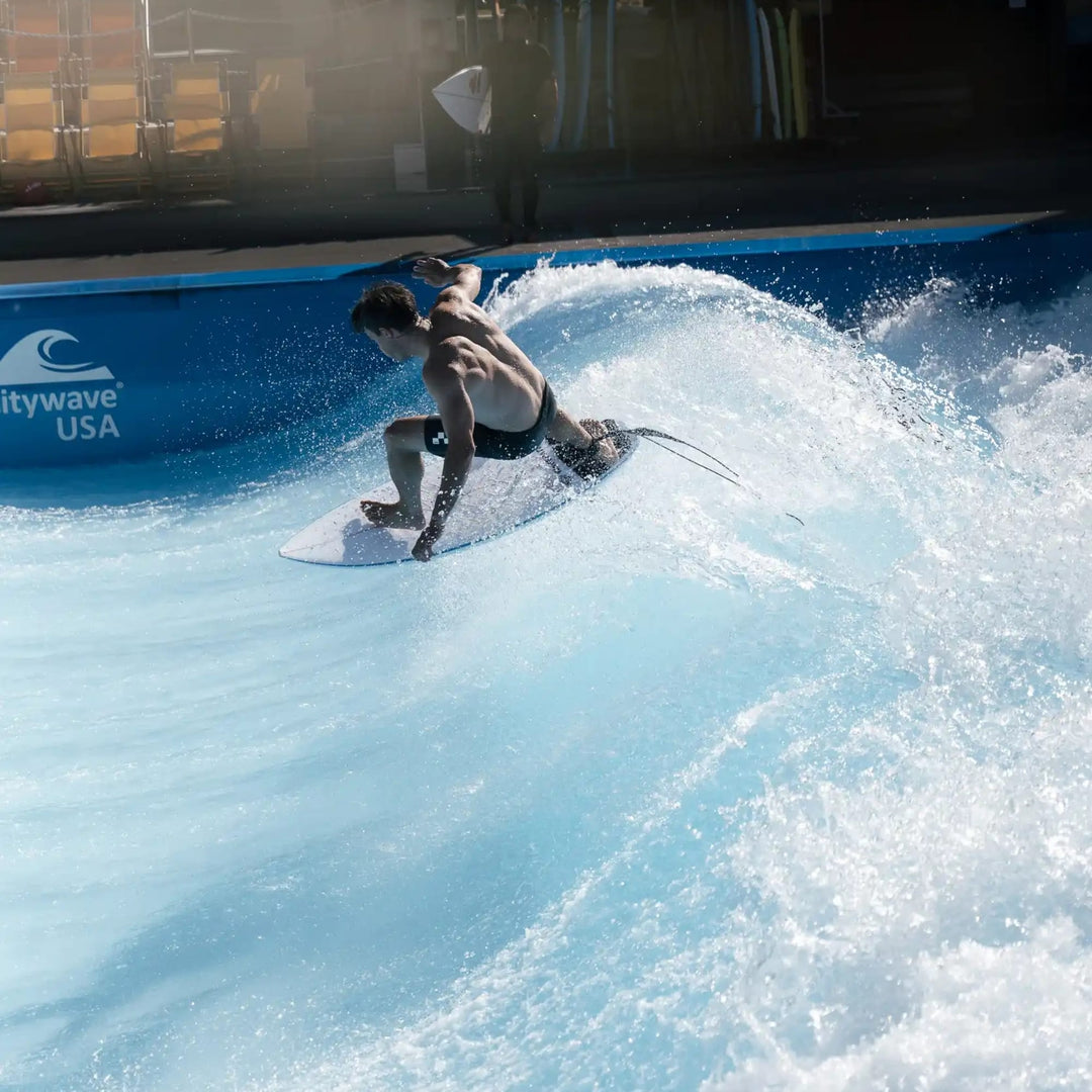 Eine Person in Badehose surft drinnen auf einer künstlichen Welle und stützt sich auf ein NOA Surfboards - CSQ von NOA, während das Wasser um sie herum spritzt. Das Wellenbad trägt die Aufschrift "citywave USA", im Hintergrund sind weitere Surfboards zu sehen.