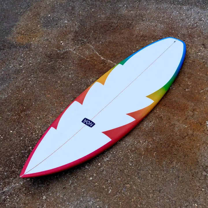 Ein NOA Surfboards - CSQ Fluss-Surfbrett mit weißer Mitte und einem von Blau zu Rot übergehenden Rand ruht auf rauem braunem Beton. Das NOA-Logo in der Mitte suggeriert Kontrolle und Geschwindigkeit für aufregende Fluss-Surfabenteuer.